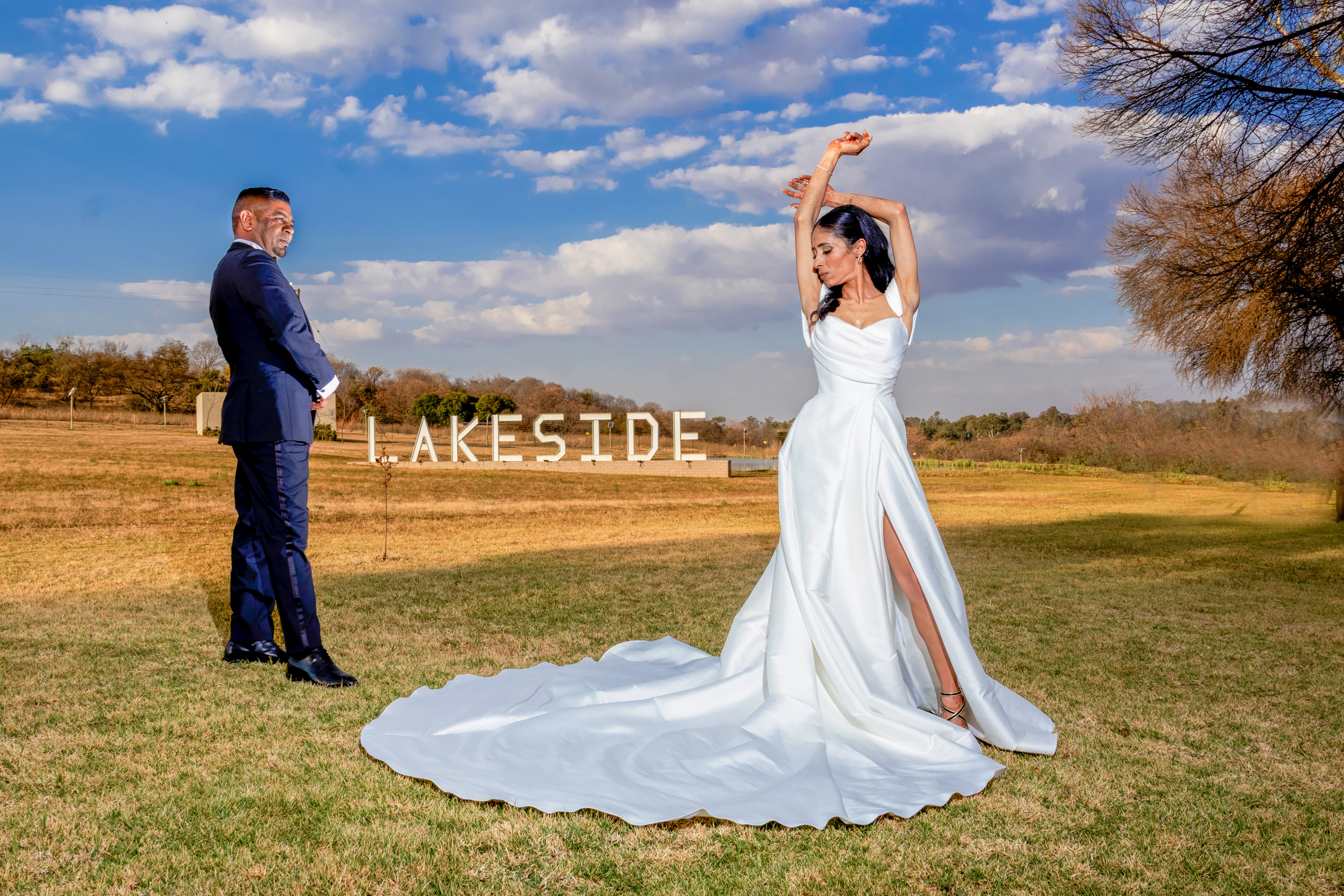 Bride posing elegantly with arms raised as the groom looks on, Lakeside sign behind