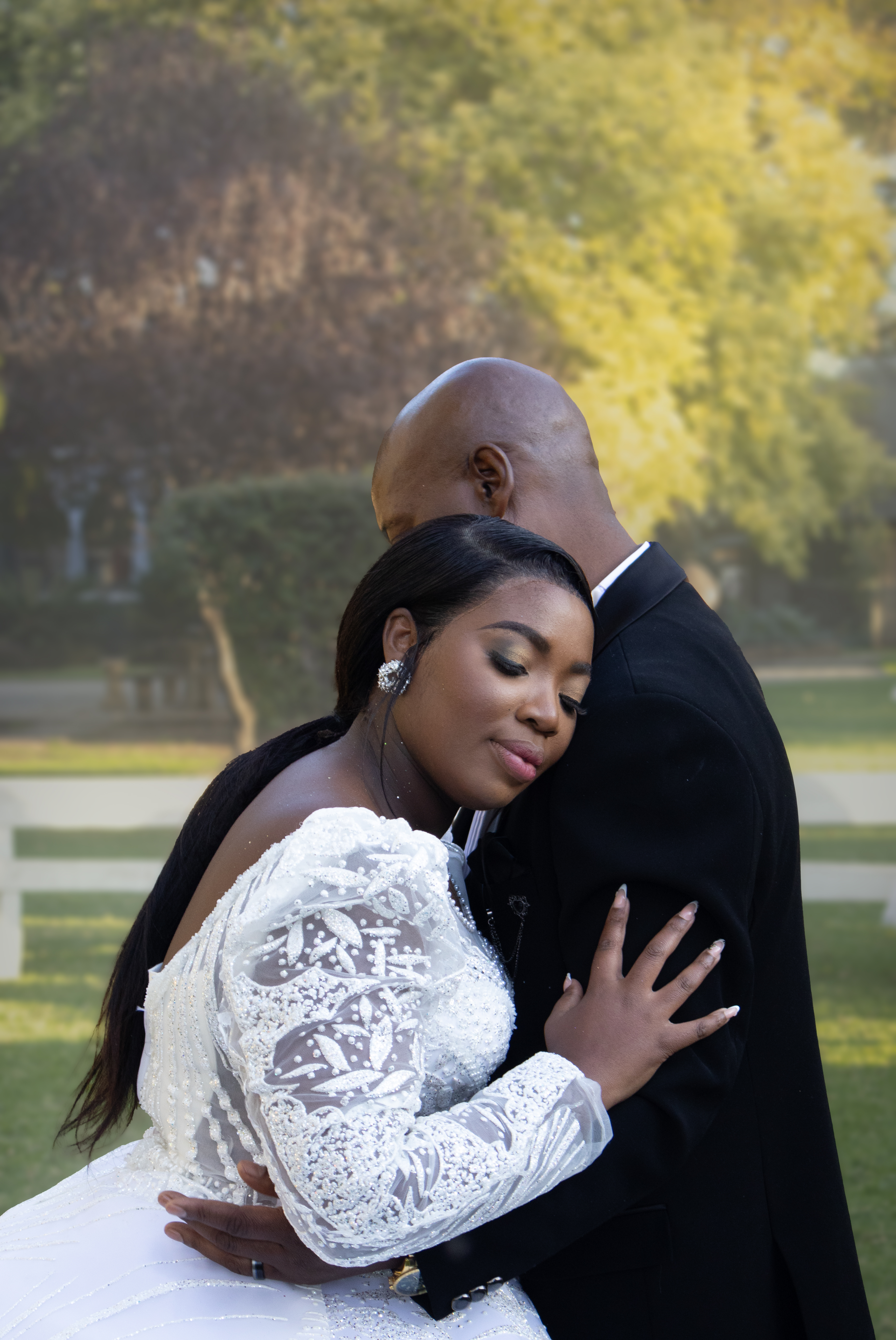 Bride in a beaded lace gown resting on the groom's shoulder in a sunlit garden
