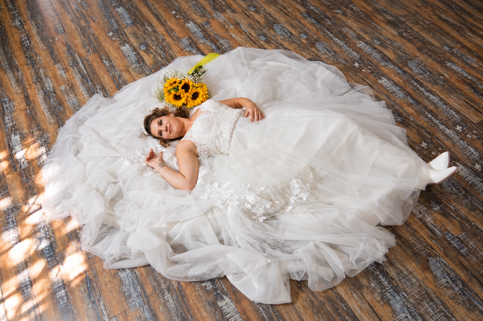Bride lying on a wooden floor with her tulle gown spread out and a sunflower bouquet