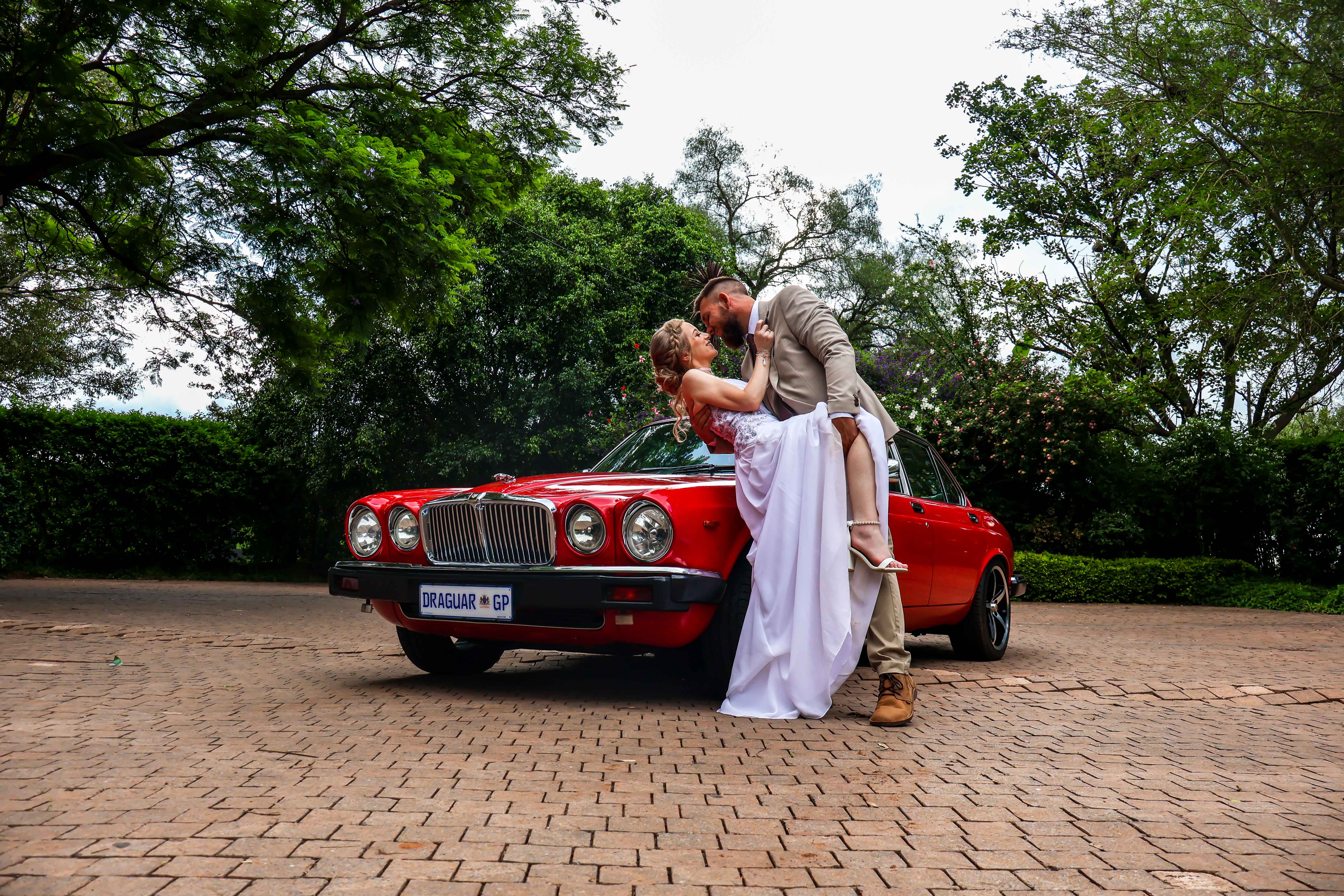 Couple sharing a dip kiss beside a classic red Jaguar