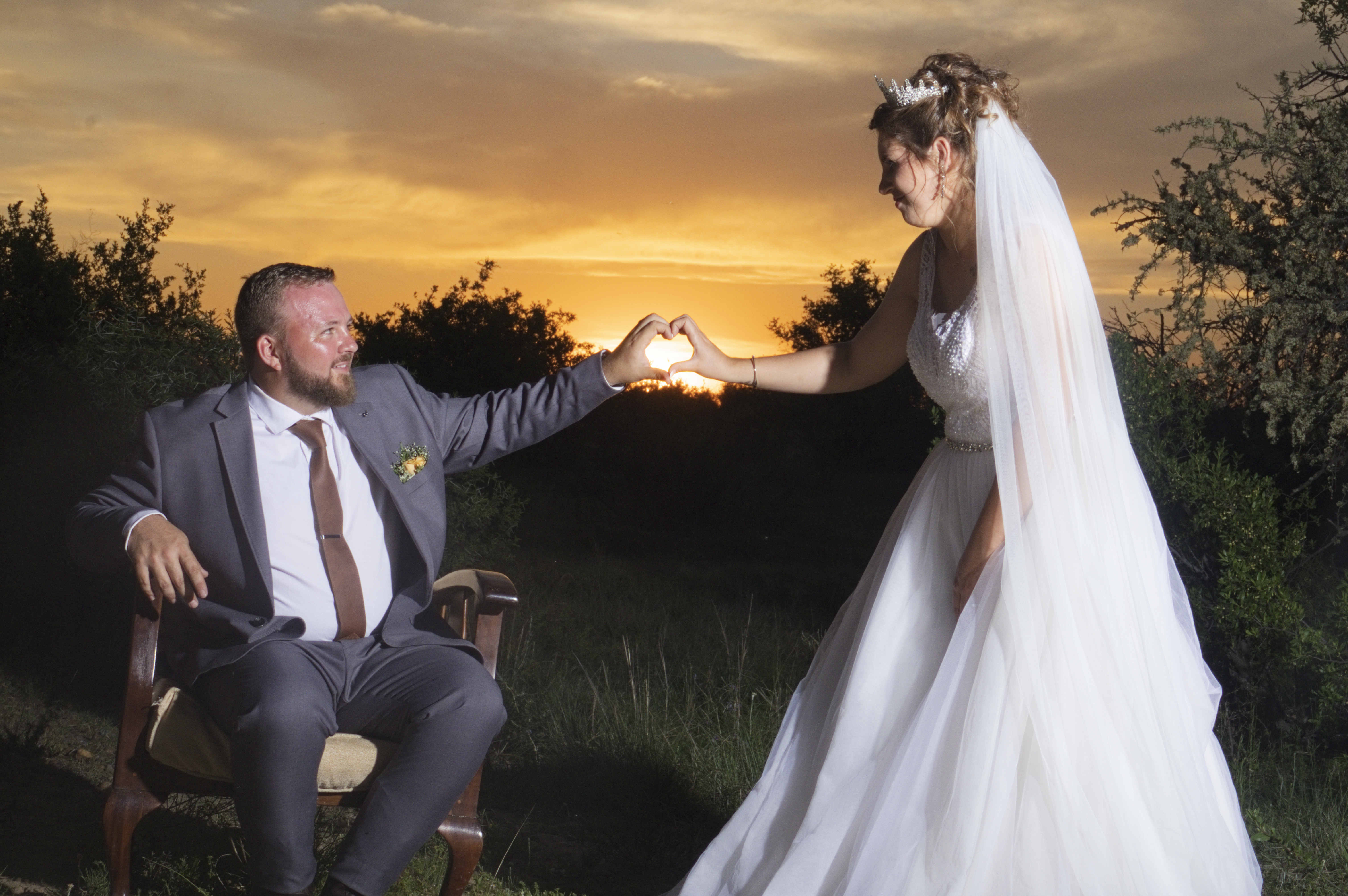 Seated groom and bride joining hands in a heart against the sunset