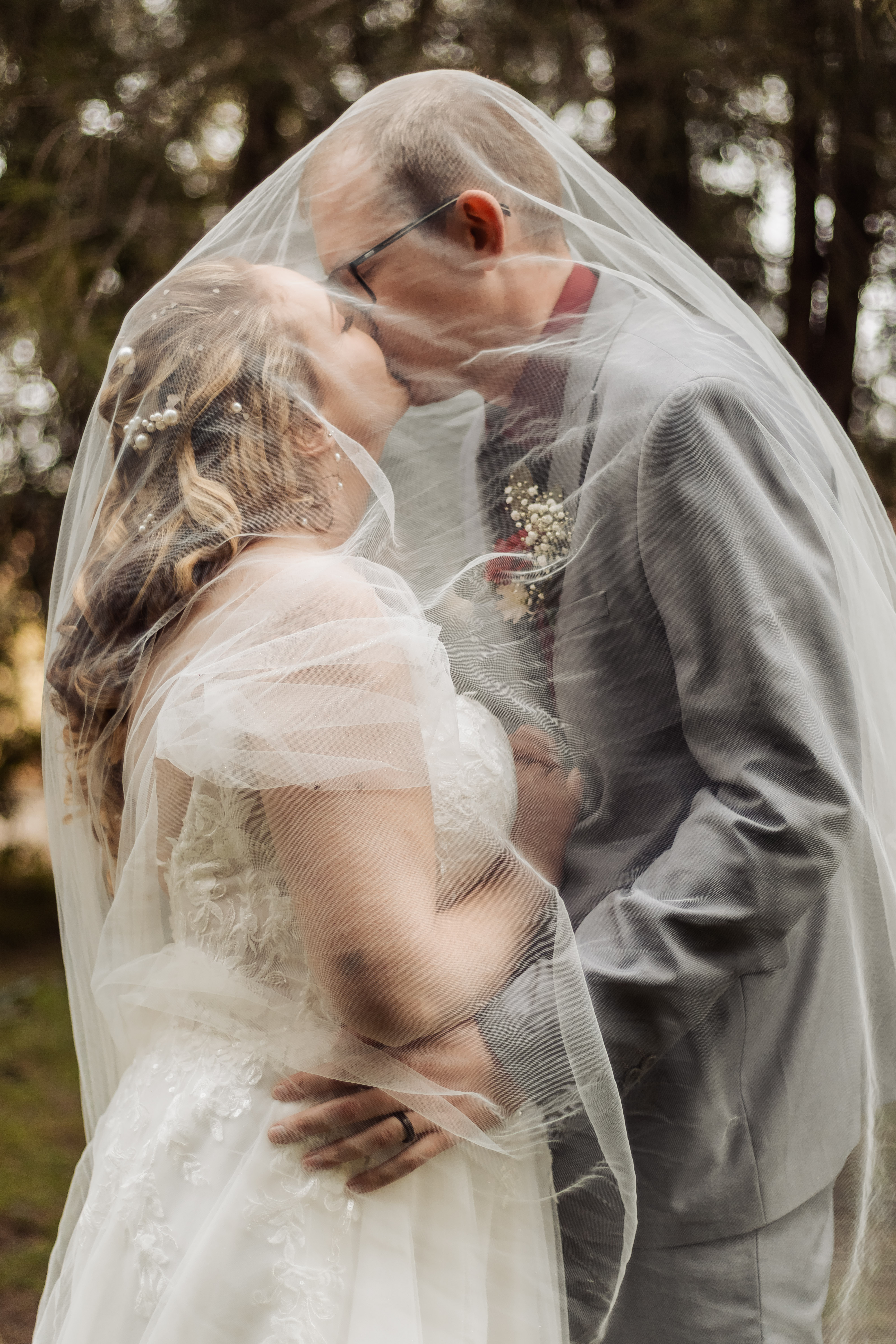 Bride and groom kissing beneath a sheer veil in a forest