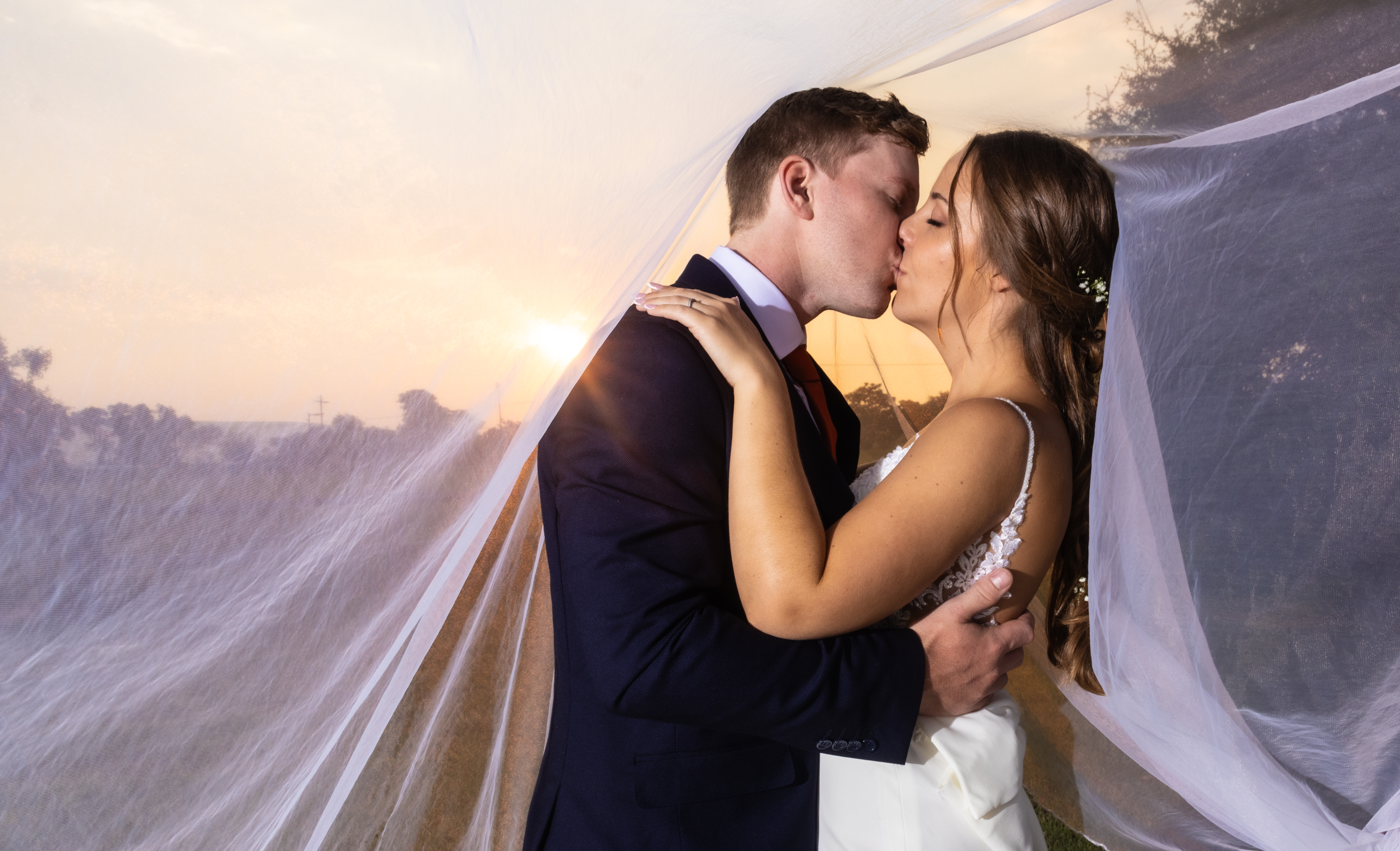 Couple kissing beneath a sheer veil as the sun sets
