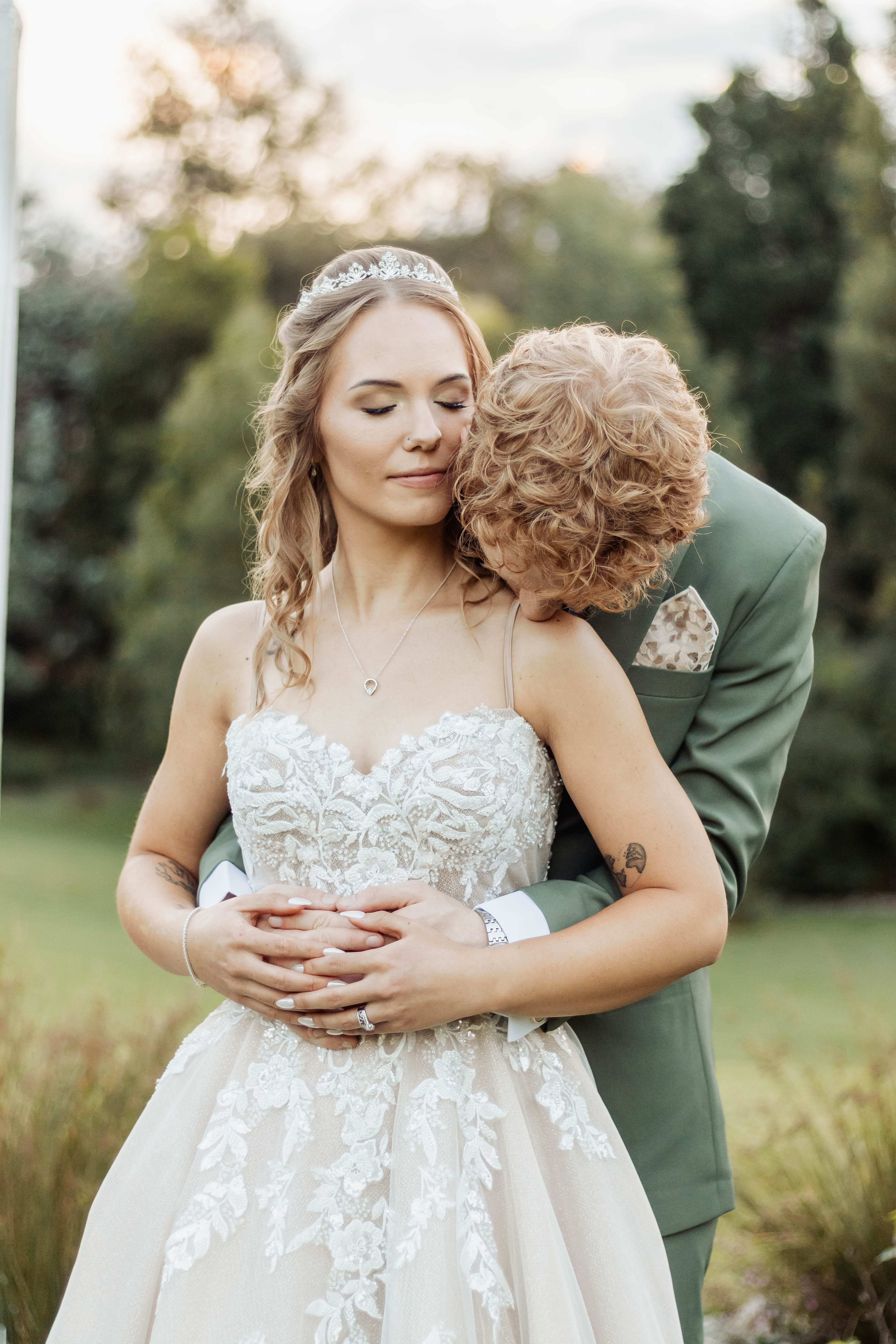 Groom in a sage suit embracing the bride from behind in a garden